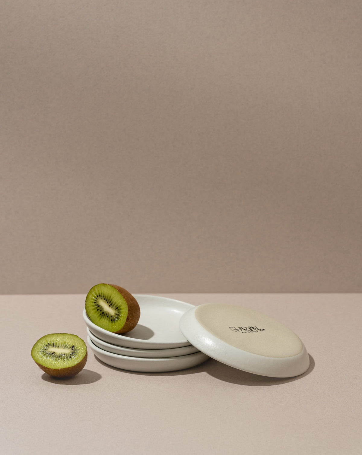 A white stoneware flat side plate with a matte finish, displayed with a halved kiwi on top, against a neutral background.
