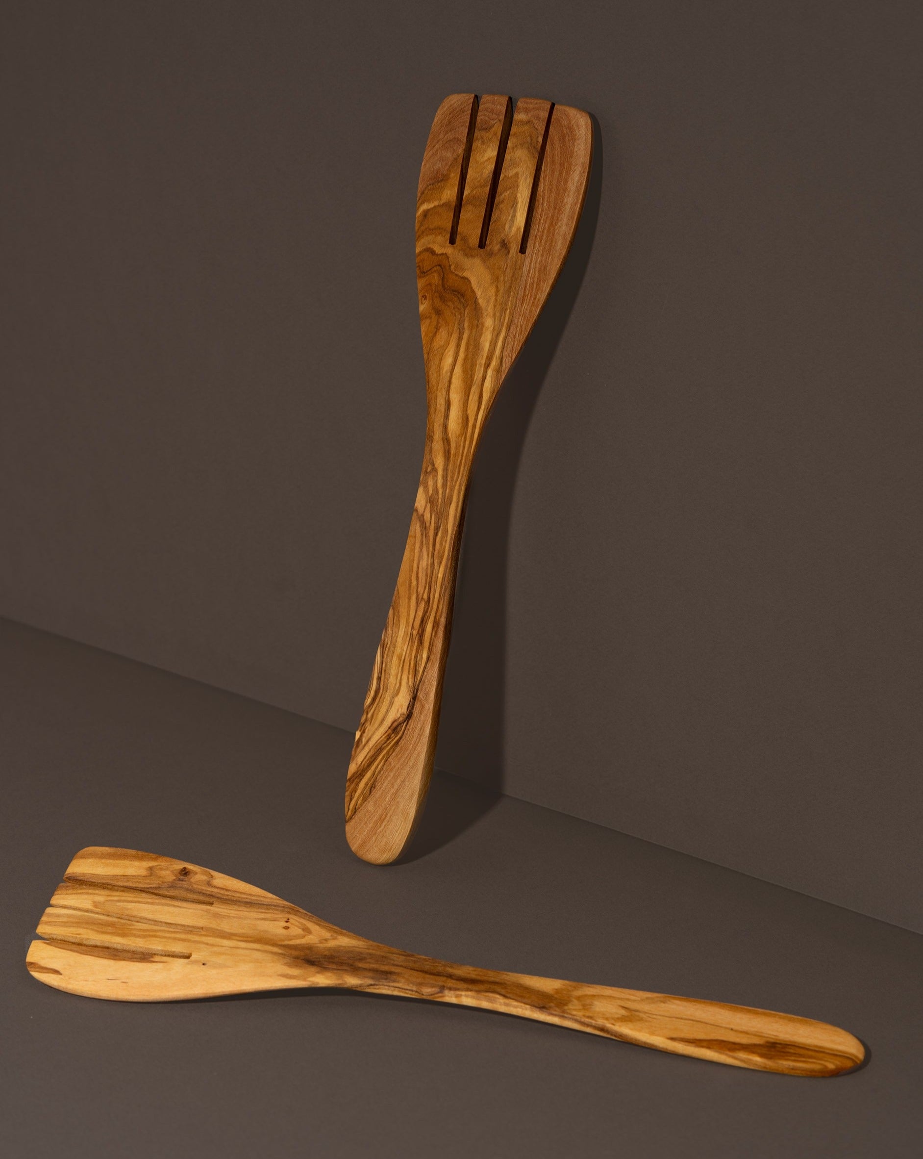 A pair of natural olive wood serving spatula forks laid on a dark surface.