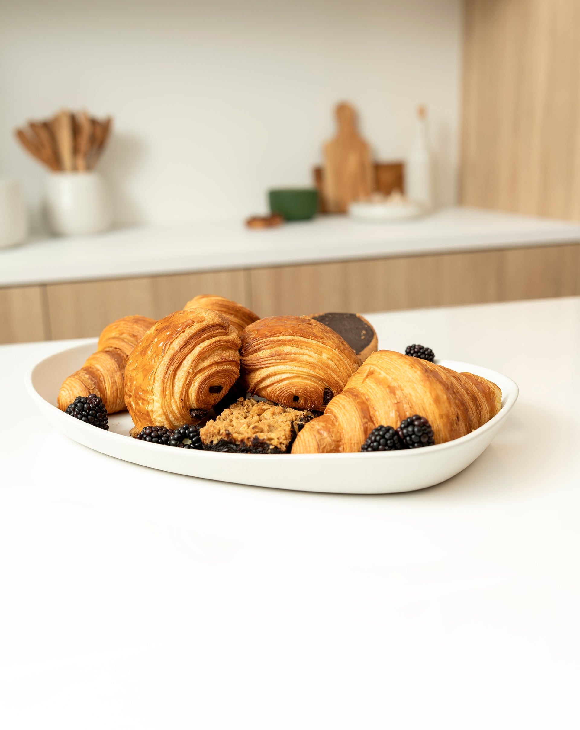 A white Stoneware platter filled with fresh croissants, a chocolate pastry, oat bars, and blackberries. The platter sits on a clean white kitchen counter, with wooden utensils and minimalist decor in the background, reflecting GHARYAN’s modern and natural aesthetic
