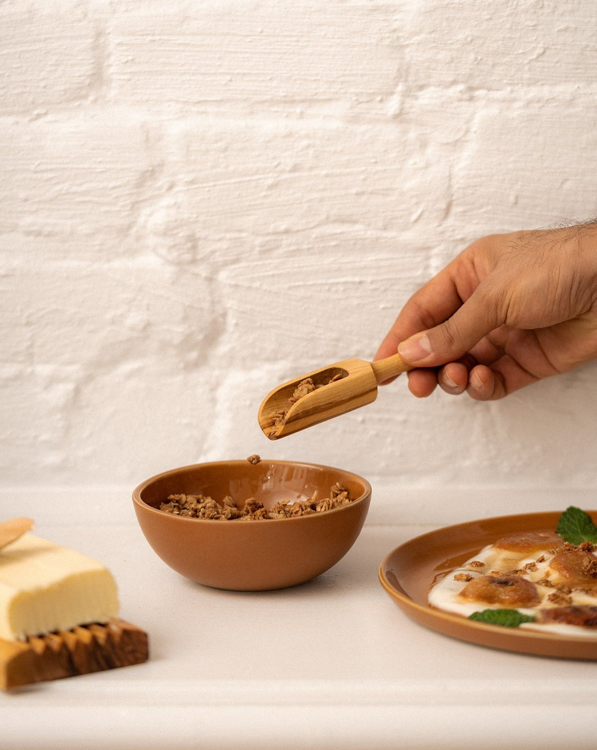 A wooden scoop resting inside a filled bowl. Beside it are a plate with food and a wooden cheese board topped with butter, accompanied by a spreader—all set against a neutral background for a cozy, rustic presentation.