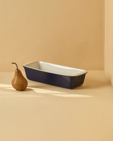 A white enameled stoneware cake and bread pan on a beige background, with two red apples placed on top of it to the side.
