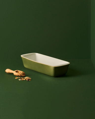A white enameled stoneware cake and bread pan on a beige background, with two red apples placed on top of it to the side.